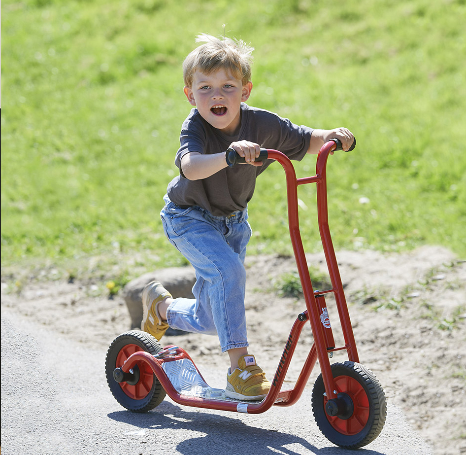 A little boy on a winter brand bright red scooter. The scooter is chunky and solid looking with solid wheels, a large standing platform and rubber grips on the red handlebars