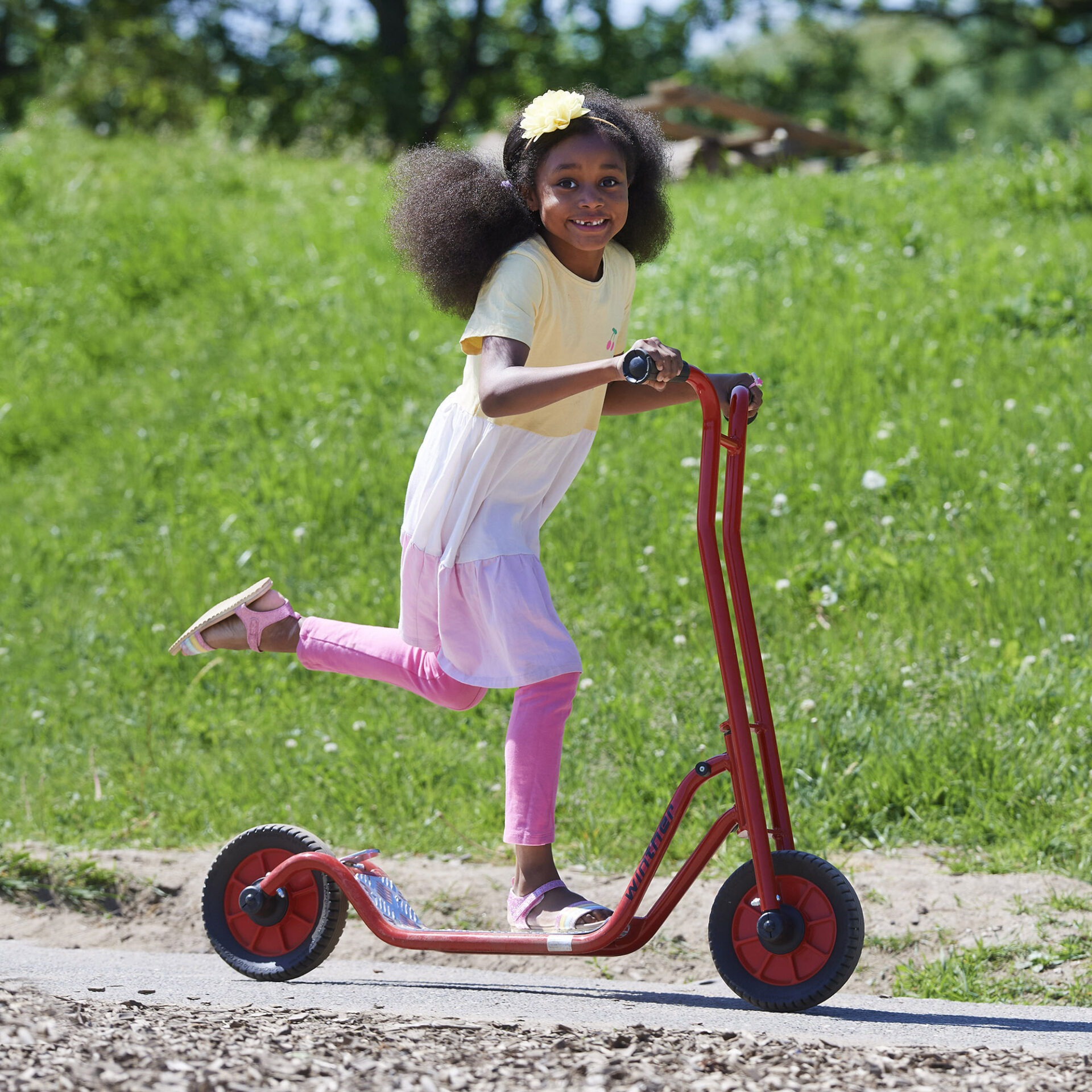 A little girl is going past at speed with a big smile riding a winther brand bright red scooter. The scooter is solid and strong looking with solid red wheels and black tyres